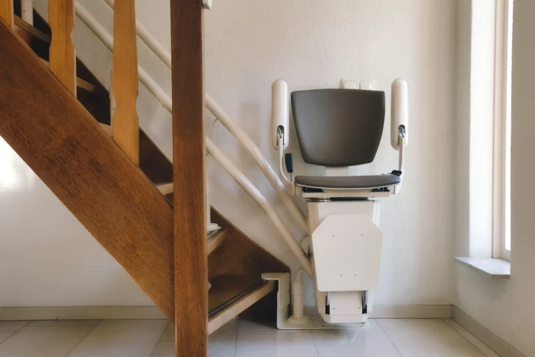 Treppenlift an einer Holztreppe in einem Wohnraum. Das Gerät ist kompakt gestaltet und ermöglicht barrierefreie Mobilität für Personen mit eingeschränkter Bewegungsfähigkeit. Es ist an einer weißen Führungsschiene entlang der Treppe befestigt und verfügt über eine ergonomische Sitzfläche mit Armlehnen.