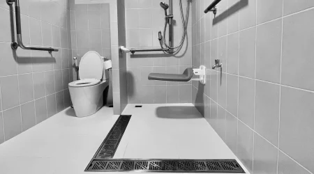 Schwarz-weiß Foto eines barrierefreien Badezimmers mit bodengleicher Dusche. Die Dusche ist mit einem klappbaren Duschsitz, einer Handbrause und mehreren Haltegriffen ausgestattet. Eine Toilette mit Haltegriffen befindet sich links daneben. Der Boden hat ein breites, integriertes Ablaufsystem, und an der Wand ist ein Fenster für natürliches Licht.