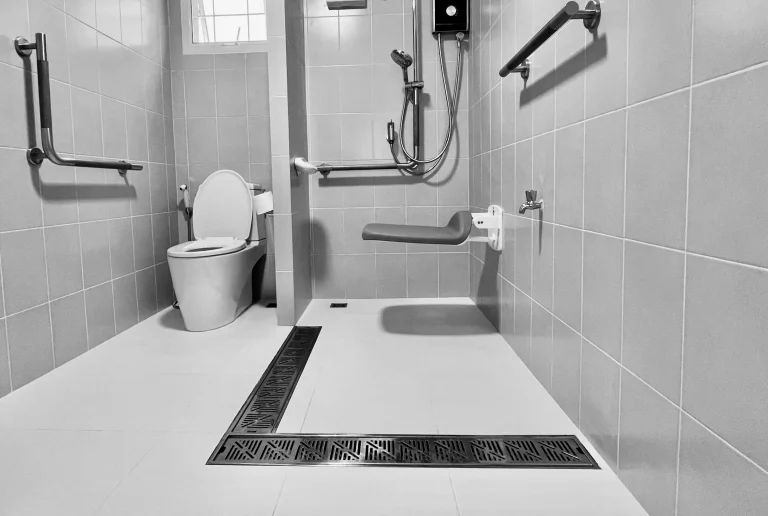 Schwarz-weiß Foto eines barrierefreien Badezimmers mit bodengleicher Dusche. Die Dusche ist mit einem klappbaren Duschsitz, einer Handbrause und mehreren Haltegriffen ausgestattet. Eine Toilette mit Haltegriffen befindet sich links daneben. Der Boden hat ein breites, integriertes Ablaufsystem, und an der Wand ist ein Fenster für natürliches Licht.