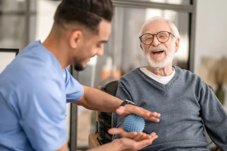 Älterer Mann mit Brille und grauem Pullover sitzt auf einem Stuhl und hält einen blauen Massageball in der Hand. Ein Pfleger in blauer medizinischer Kleidung unterstützt ihn dabei. Im Hintergrund sind große Fenster und eine moderne Einrichtung sichtbar.