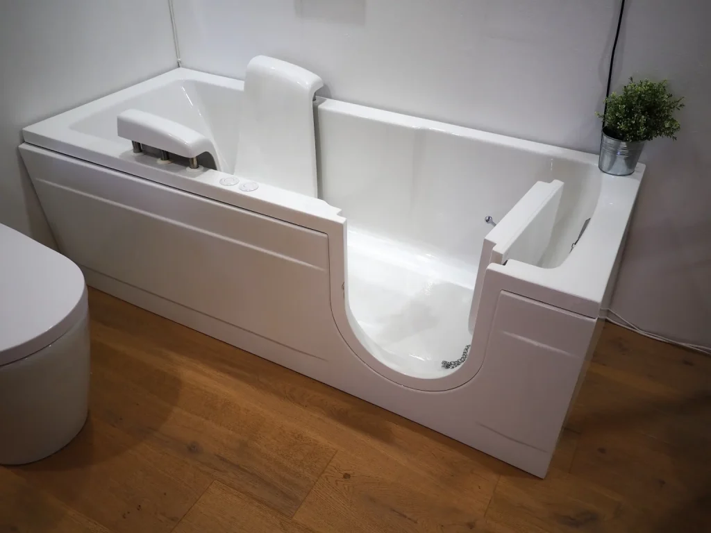 Sitzbadewanne mit seitlicher Tür für barrierefreien Zugang in einem modernen Badezimmer. Die Badewanne ist weiß, hat eine integrierte Rückenlehne und eine Armstütze mit Wasserhahn. Der Boden besteht aus Holzoptik, daneben steht eine Toilette. An der Wand ist eine kleine grüne Pflanze in einem Metalltopf platziert.