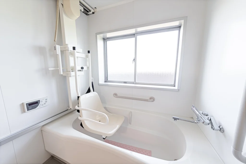 Barrierefreie Badewanne mit integriertem elektrischen Hebesitz in einem hellen Badezimmer. Der weiße Hebesitz mit Armlehnen ist an einer Deckenhalterung befestigt und ermöglicht einen sicheren Ein- und Ausstieg. Die Badewanne ist mit Haltegriffen ausgestattet, und an der Wand befindet sich ein modernes Bedienpanel. Ein großes Fenster sorgt für natürliches Licht.