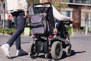 Person in einem elektrischen Rollstuhl mit montiertem Rucksack fährt auf einem gepflasterten Gehweg in urbaner Umgebung. Eine weitere Person geht daneben.