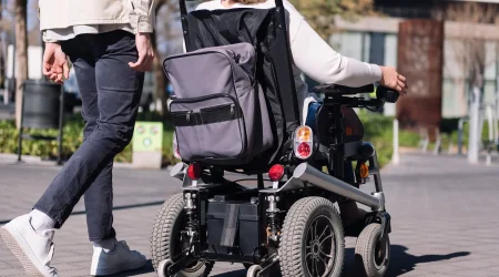 Person in einem elektrischen Rollstuhl mit montiertem Rucksack fährt auf einem gepflasterten Gehweg in urbaner Umgebung. Eine weitere Person geht daneben.