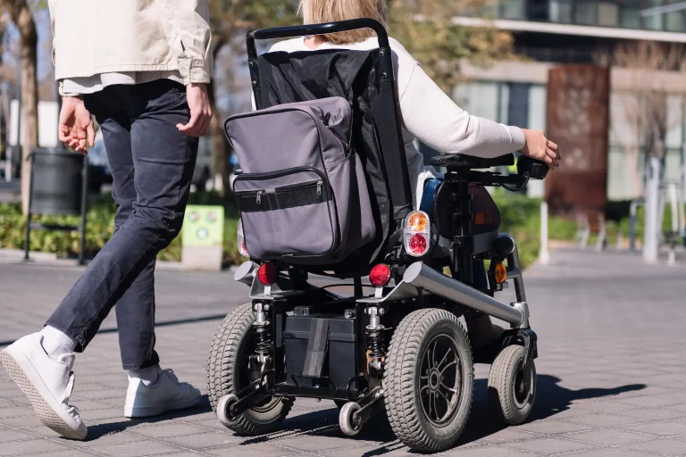 Person in einem elektrischen Rollstuhl mit montiertem Rucksack fährt auf einem gepflasterten Gehweg in urbaner Umgebung. Eine weitere Person geht daneben.
