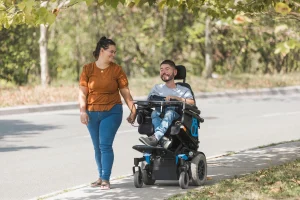 Ein Mann im elektrischen Rollstuhl fährt lächelnd auf einem Gehweg, während er die Hand einer Frau hält, die neben ihm geht. Beide wirken fröhlich und genießen gemeinsam einen sonnigen Tag im Freien. Im Hintergrund sind Bäume und eine Straße zu sehen.