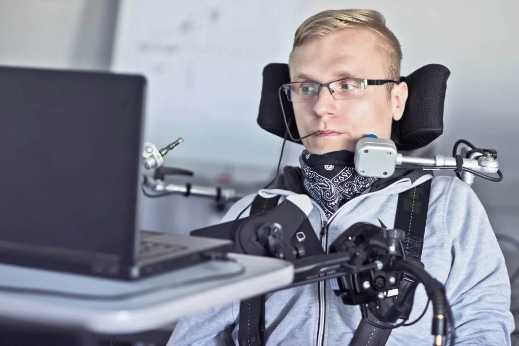 Ein junger Mann im Elektrorollstuhl nutzt einen Laptop. Er steuert den Computer über ein Mundstück und wird von einem komplexen technischen Assistenzsystem unterstützt. Der Fokus liegt auf der Barrierefreiheit und der Nutzung moderner Hilfsmittel für Menschen mit körperlicher Behinderung.