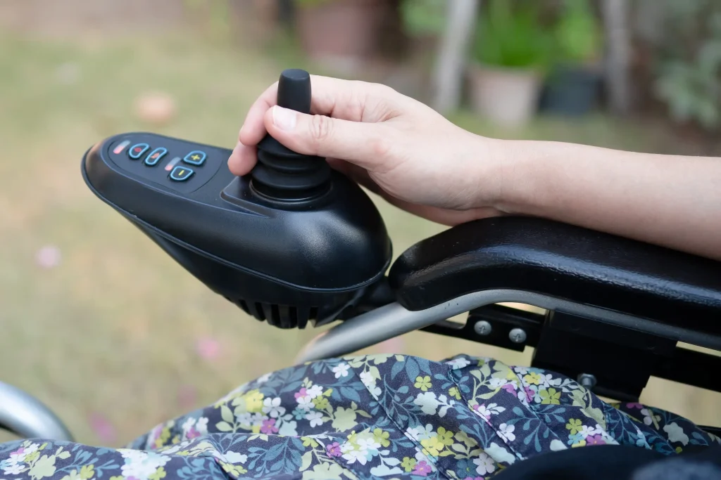 Hand einer Person hält den Joystick eines Elektrorollstuhls. Die Steuerkonsole ist mit mehreren farbigen Tasten versehen, der Rollstuhl hat eine schwarze Armlehne. Im Hintergrund ist unscharf eine Rasenfläche zu sehen.