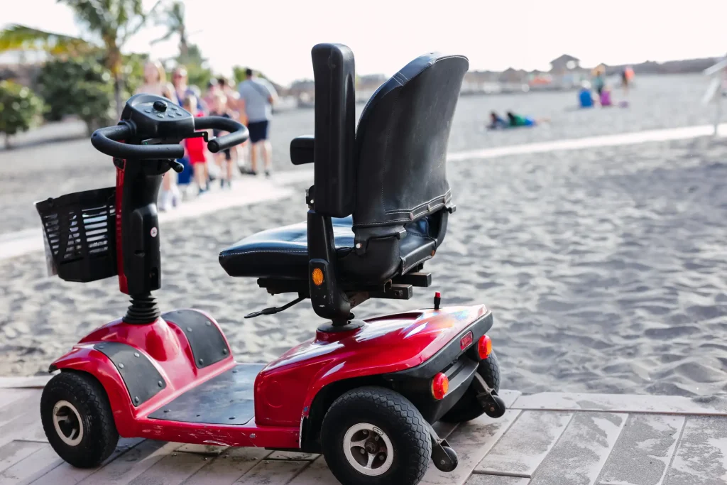 Rotes vierrädriges Elektromobil mit schwarzem Sitz, Armlehnen und Lenker mit Korb steht auf einem Holzweg am Sandstrand. Im Hintergrund gehen mehrere Personen auf dem Steg, einige sitzen oder liegen im Sand, Palmen und Gebäude sind in der Ferne sichtbar.