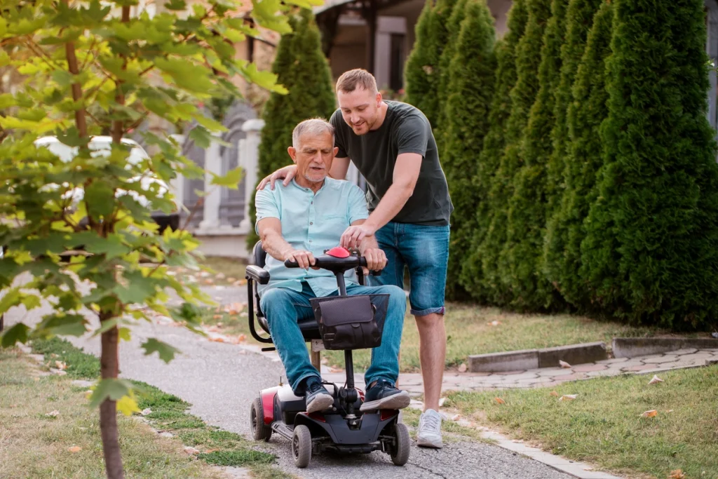 Junger Mann begleitet älteren Mann, welcher in einem Elektromobil auf einem Gehwg fährt.