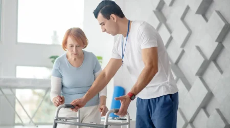 Ein Pfleger unterstützt eine ältere Frau mit rotblondem Haar beim Gehen mit einem Rollator in einer hellen, modernen Rehabilitationsumgebung. Der Pfleger zeigt ihr ruhig und konzentriert, wie sie den Gehwagen sicher benutzt.