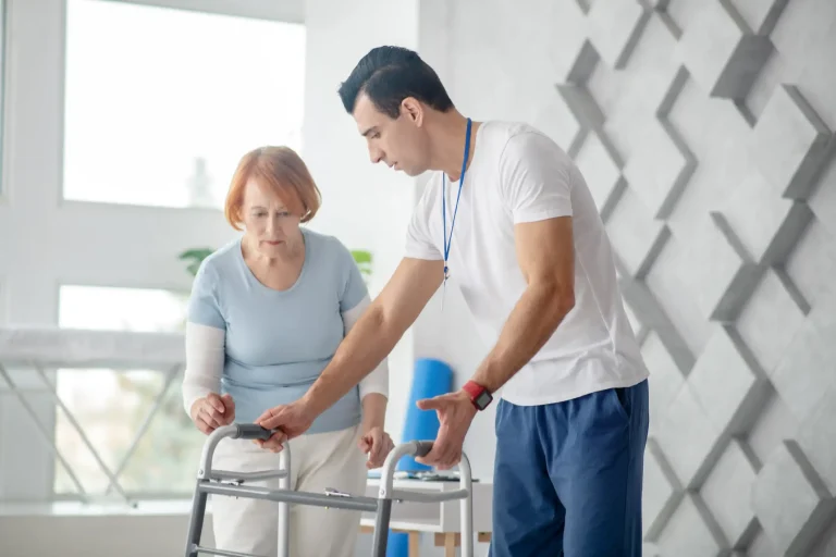 Ein Pfleger unterstützt eine ältere Frau mit rotblondem Haar beim Gehen mit einem Rollator in einer hellen, modernen Rehabilitationsumgebung. Der Pfleger zeigt ihr ruhig und konzentriert, wie sie den Gehwagen sicher benutzt.