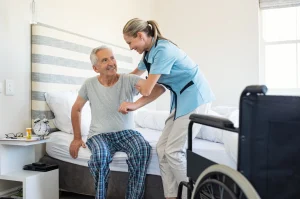 Pflegekraft hilft älterem Herrn aus dem Bett in den Rollstuhl