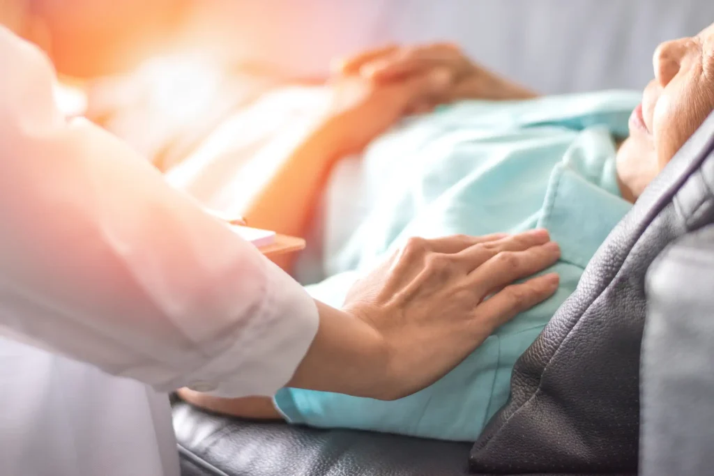 Pflegekraft legt beruhigend eine Hand auf die Schulter einer älteren Frau, die in einem Krankenbett liegt. Die Frau trägt ein hellblaues Hemd und ruht mit gefalteten Händen.