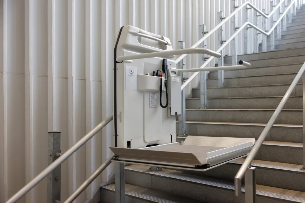 Ein leerer Plattformlift für Rollstuhlfahrer ist an einem Treppengeländer außen installiert. Der Lift ist hochgeklappt und befindet sich am unteren Ende einer langen Treppe mit Handläufen. Die Anlage ist für barrierefreien Zugang in öffentlichen oder mehrstöckigen Gebäuden vorgesehen.