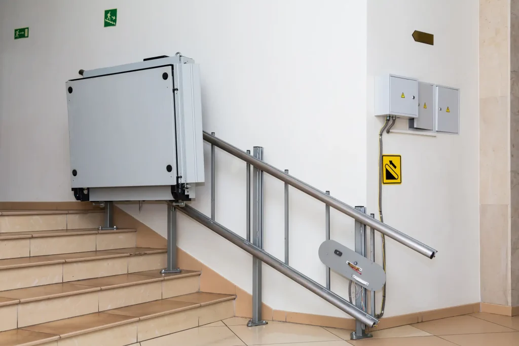 Ein zusammengeklappter Plattformlift ist innen an einem Treppengeländer montiert. Die Anlage dient dem barrierefreien Zugang für Rollstuhlfahrer. Der Lift ist an einer gefliesten Treppe installiert, daneben befinden sich elektrische Schaltschränke und ein Warnschild an der Wand.