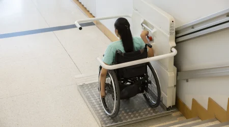 Eine Frau im Rollstuhl nutzt einen Plattformlift, um eine Treppe hinunterzufahren. Der Lift ist an einem Schienensystem an der Wand befestigt und ermöglicht barrierefreies Überwinden von Höhenunterschieden in öffentlichen Gebäuden.