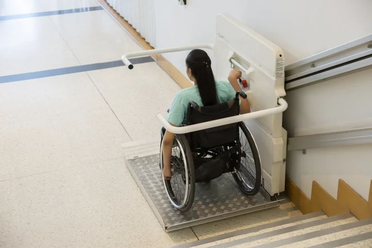 Eine Frau im Rollstuhl nutzt einen Plattformlift, um eine Treppe hinunterzufahren. Der Lift ist an einem Schienensystem an der Wand befestigt und ermöglicht barrierefreies Überwinden von Höhenunterschieden in öffentlichen Gebäuden.
