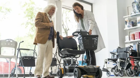 Älterer Mann in einem Sanitätshaus lässt sich von einer Mitarbeiterin ein Elektromobil zeigen. Im Hintergrund sind Rollatoren und Rollstühle ausgestellt.
