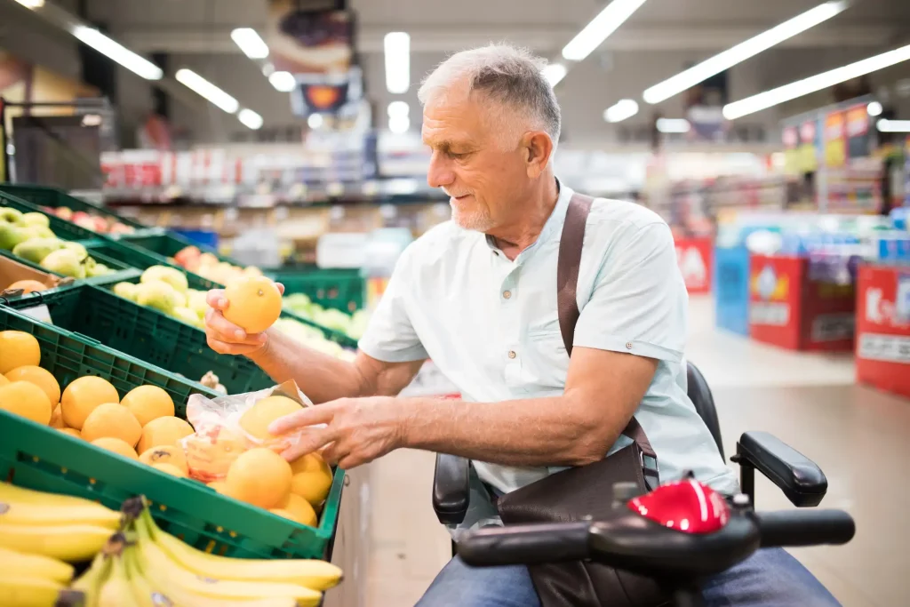 Älterer Mann auf einem Elektromobil wählt im Supermarkt Orangen aus einer grünen Auslage mit verschiedenen Obstsorten.