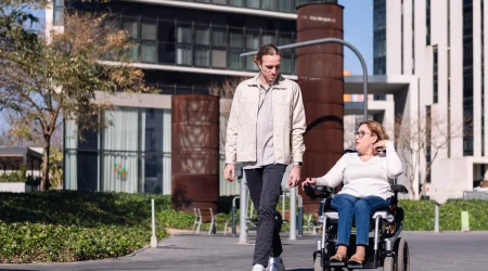Eine Frau fährt in einem faltbaren Elektrorollstuhl neben einem Mann, der zu Fuß geht. Sie unterhalten sich auf einem barrierefreien Weg in einer modernen, urbanen Umgebung mit Bürogebäuden im Hintergrund.