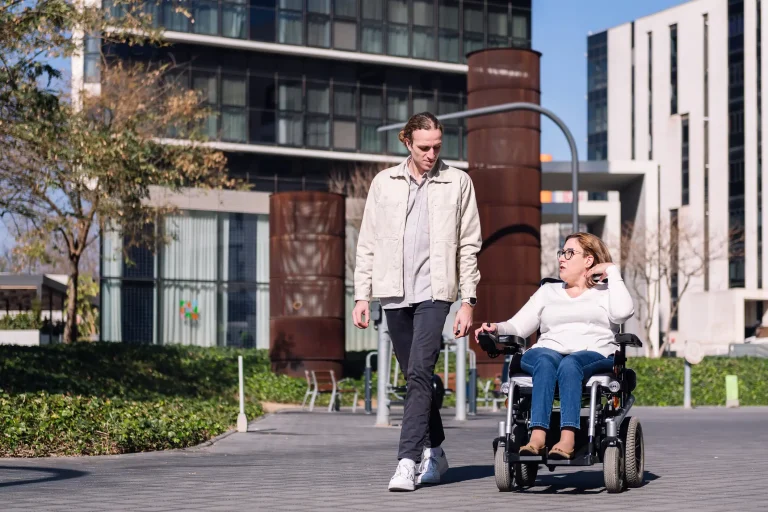 Eine Frau fährt in einem faltbaren Elektrorollstuhl neben einem Mann, der zu Fuß geht. Sie unterhalten sich auf einem barrierefreien Weg in einer modernen, urbanen Umgebung mit Bürogebäuden im Hintergrund.