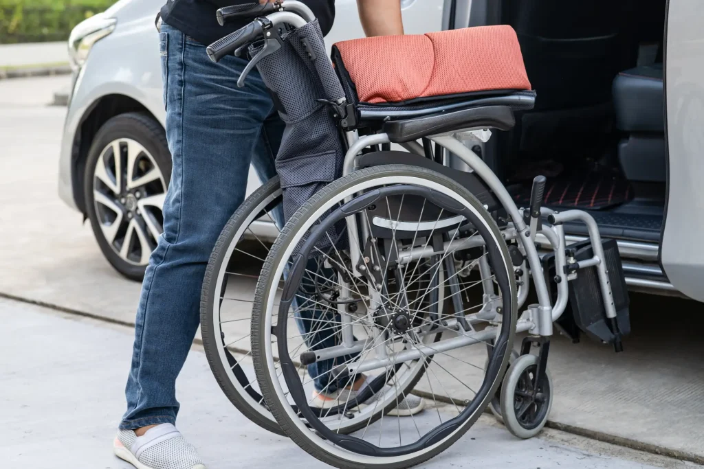 Eine Person verstaut einen zusammengeklappten, manuellen Rollstuhl mit rotem Sitzkissen in ein Auto. Das Fahrzeug steht geöffnet auf einem Parkplatz.