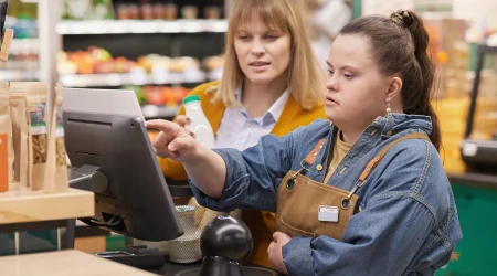 Ein Mädchen mit Behinderung wird im Supermarkt an der Registrierkasse eingearbeitet und erhält Unterstützung beim selbstbestimmten Lernen in einem inklusiven Arbeitsumfeld, das Teilhabe am Berufsleben und Chancengleichheit fördert.