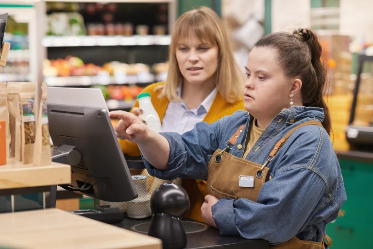 Ein Mädchen mit Behinderung wird im Supermarkt an der Registrierkasse eingearbeitet und erhält Unterstützung beim selbstbestimmten Lernen in einem inklusiven Arbeitsumfeld, das Teilhabe am Berufsleben und Chancengleichheit fördert.