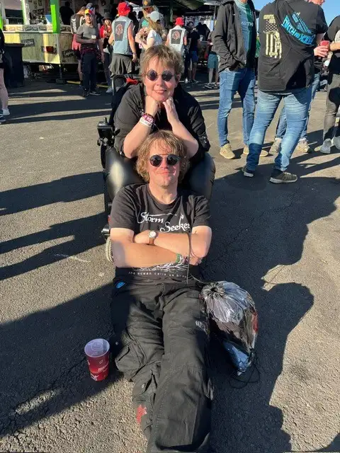 Zwei Personen bei einem Festival, hinten sitzt Petra, unsere Referenzkundin, in einem faltbaren Elektrorollstuhl. Sie sitzt mit Sonnenbrille im Rollstul, vorne sitzt ein Mann mit verschränkten Armen auf dem Boden, neben ihm stehen ein Becher und ein Rucksack.