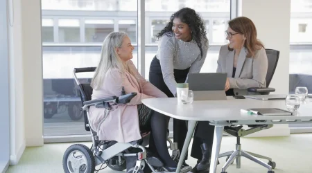 rei Frauen in einem modernen Besprechungsraum. Eine Frau sitzt in einem faltbaren Elektrorollstuhl am Tisch, daneben zwei Kolleginnen im Gespräch mit Laptop und Unterlagen.