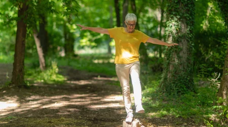 Eine ältere Frau balanciert im Wald auf einem Baumstamm.
