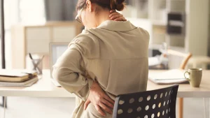 Eine Frau sitzt auf einem Bürostuhl und hält sich mit beiden Händen den Rücken.