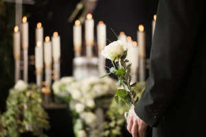 Ein Mann steht vor einem Sarg mit einer weißen Rose in den Händen. Im Hintergrund stehen brennende weiße Kerzen.