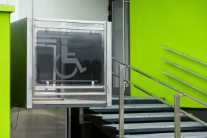 Ein elektrisch betriebener Hublift mit transparenter Umrandung befindet sich neben einer Treppe an einem Gebäude mit grün gestrichener Außenwand. Auf der Front des Lifts ist ein Rollstuhlsymbol abgebildet. Der Lift dient dem barrierefreien Zugang zum erhöhten Eingangsbereich.