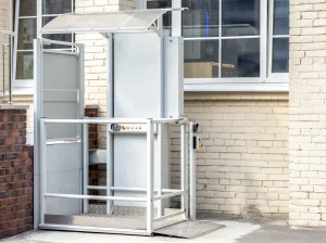 Ein außen angebrachter Treppenlift mit Plattform ist an der Fassade eines Gebäudes montiert. Der Außenlift verfügt über ein umlaufendes Schutzgeländer, ein Bedienfeld mit mehreren Tasten und ein transparentes Schutzdach. Eine Rampe mit rutschfester Metalloberfläche ermöglicht den ebenerdigen Zugang zur Plattform.