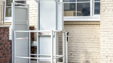 Ein außen angebrachter Treppenlift mit Plattform ist an der Fassade eines Gebäudes montiert. Der Außenlift verfügt über ein umlaufendes Schutzgeländer, ein Bedienfeld mit mehreren Tasten und ein transparentes Schutzdach. Eine Rampe mit rutschfester Metalloberfläche ermöglicht den ebenerdigen Zugang zur Plattform.