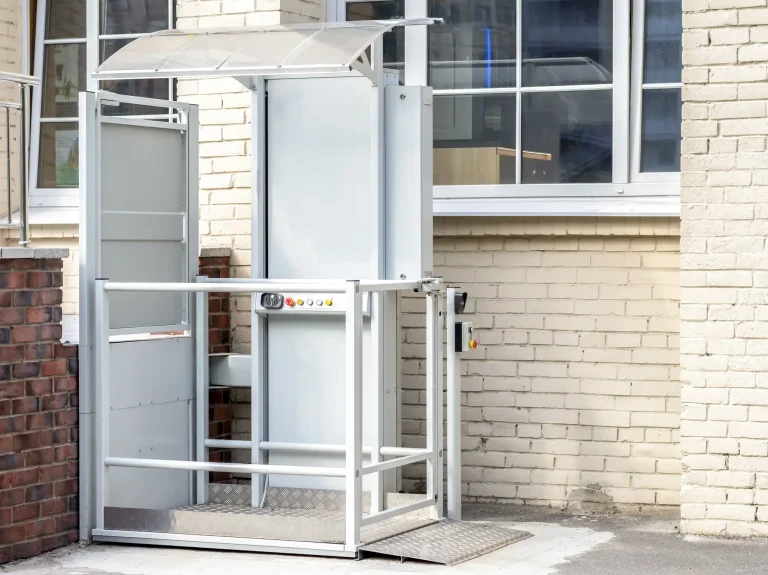 Ein außen angebrachter Treppenlift mit Plattform ist an der Fassade eines Gebäudes montiert. Der Außenlift verfügt über ein umlaufendes Schutzgeländer, ein Bedienfeld mit mehreren Tasten und ein transparentes Schutzdach. Eine Rampe mit rutschfester Metalloberfläche ermöglicht den ebenerdigen Zugang zur Plattform.