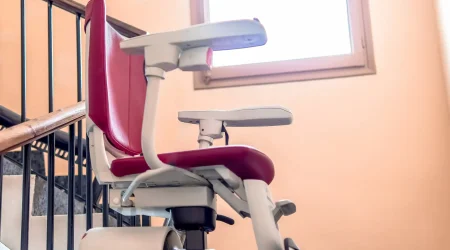 Ein Treppenlift mit rotem Sitz und klappbaren Armlehnen ist an einem Handlauf im Treppenhaus eines Mehrfamilienhauses montiert. Der Lift verläuft entlang der Treppe und ermöglicht auch Mieterinnen und Mietern mit eingeschränkter Mobilität den Zugang zu oberen Etagen.