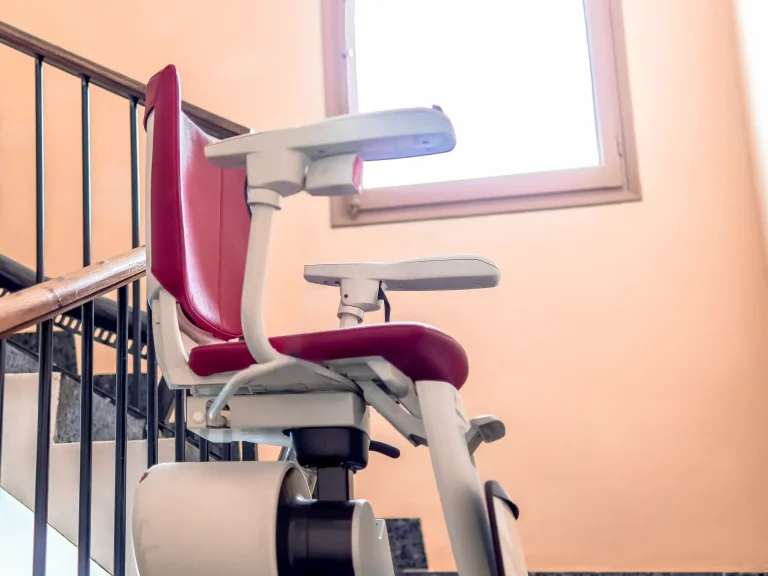 Ein Treppenlift mit rotem Sitz und klappbaren Armlehnen ist an einem Handlauf im Treppenhaus eines Mehrfamilienhauses montiert. Der Lift verläuft entlang der Treppe und ermöglicht auch Mieterinnen und Mietern mit eingeschränkter Mobilität den Zugang zu oberen Etagen.