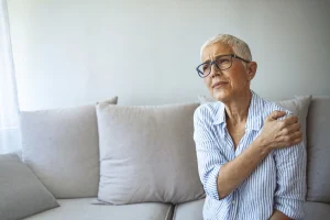 Eine ältere Frau mit kurzen grauen Haaren, sitzt auf der Couch und hält sich linke Schulter mit der rechten Hand.