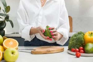Eine Frau steht in einer Küche und dreht die Hälften einer geteilten Avocado auseinander. Auf der Arbeitsfläche liegt weiteres Gemüse herum.