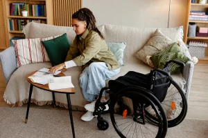 Frau sitzt auf Sofa neben einem Rollstuhl und sortiert Vertragspapiere ihrer Zusatzversicherung.