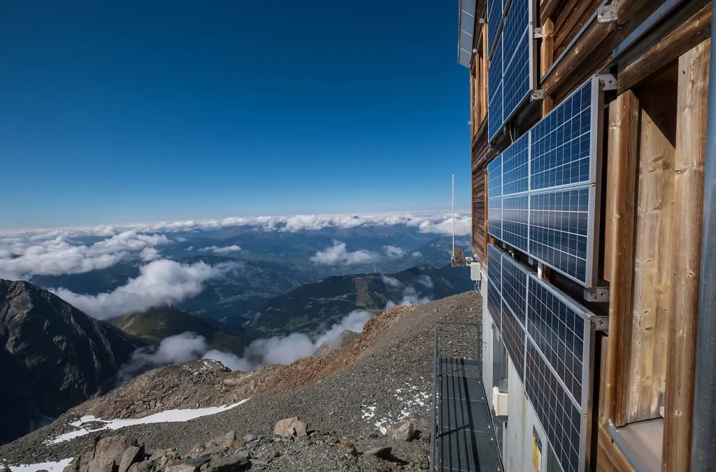 Fassadenmontierte Photovoltaikmodule einer autarken Inselanlage an einer abgelegenen Berghütte, mit Blick über umliegende Berglandschaft und Wolkenmeer.