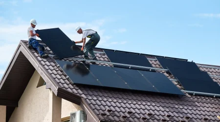 Zwei Solarteure montieren Photovoltaikmodule auf einem geneigten Ziegeldach eines Einfamilienhauses unter freiem Himmel.