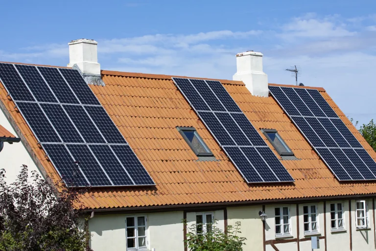 Mehrere Photovoltaikmodule sind auf einem geneigten Ziegeldach eines Wohnhauses installiert. Die Solarmodule sind gleichmäßig verteilt und um Dachfenster sowie Schornsteine herum angeordnet.