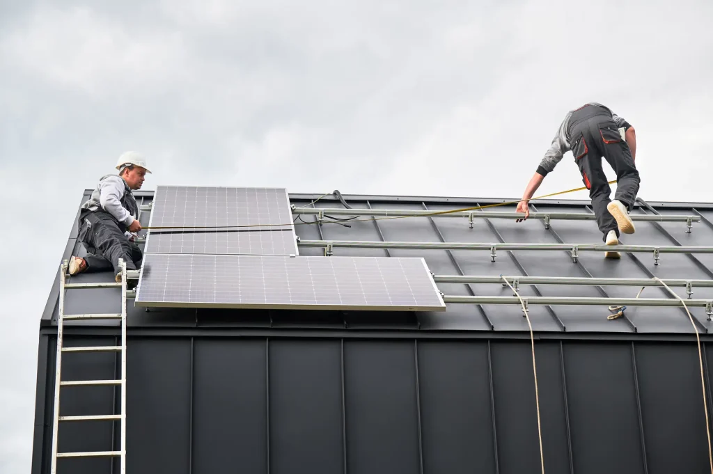 Zwei Solarteure installieren Photovoltaikmodule auf einem Metalldach. Ein Handwerker steht auf einer Leiter und richtet ein Solarmodul aus, während der andere auf dem Dach Montageschienen und Kabel befestigt.