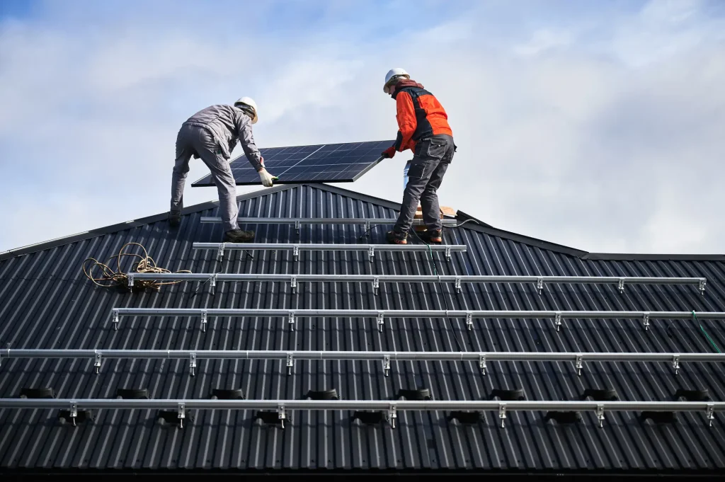 Zwei Solarteure mit Schutzhelmen platzieren ein Photovoltaik-Modul auf einem dunklen Metalldach, auf dem bereits mehrere Reihen von Montageschienen befestigt sind.