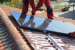 Ein Solarteur in roter Arbeitskleidung montiert ein dunkles Photovoltaik-Modul auf einem Ziegeldach, wobei die Unterkonstruktion und Verkabelung sichtbar sind.