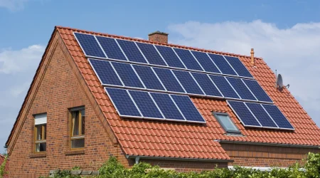 Eine großflächige Photovoltaikanlage mit 18 Solarmodulen, die auf dem roten Ziegeldach eines modernen Backsteinhauses montiert ist, unter blauem Himmel mit leichten Wolken.